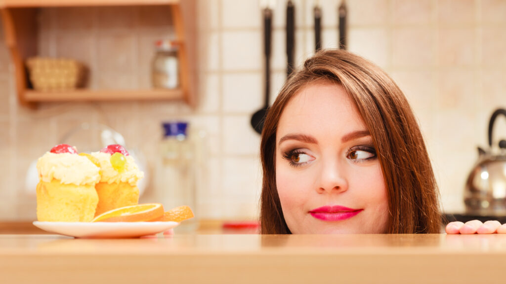 Woman hidden behind table sneaking and looking at delicious cake with sweet cream and fruits on top. Appetite and gluttony concept. Beat your food cravings
